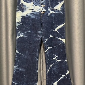 May include: Dark blue denim jeans with a white tie-dye pattern. The jeans have a button fly, front and back pockets, and a flared leg. The jeans are hanging on a hanger.