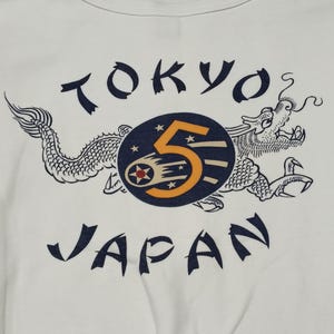 May include: An off-white t-shirt featuring a dark blue graphic print. The design includes the text TOKYO and JAPAN in a stylized font, surrounding a central dark blue circle with a large orange number 5 and a dragon illustration.