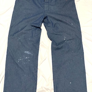 Puede incluir: Pantalones de trabajo de mezclilla azul oscuro con cierre de botón. Los pantalones tienen un diseño de pierna recta y muestran algunas salpicaduras de pintura blanca. Los pantalones están planos sobre una superficie blanca.