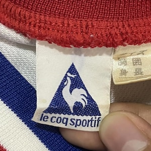 May include: Close-up of a white, red, and blue athletic shirt collar. The tag features the Le Coq Sportif logo, a blue triangle with a white rooster, and the brand name. The shirt's collar is red with white and blue stripes.