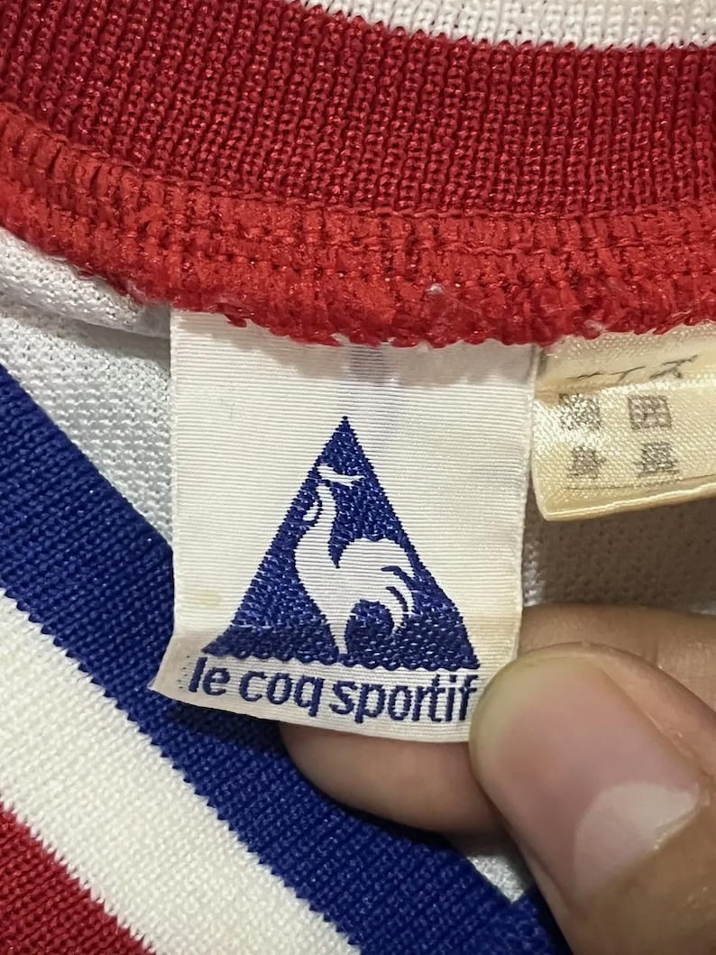 May include: Close-up of a white, red, and blue athletic shirt collar. The tag features the Le Coq Sportif logo, a blue triangle with a white rooster, and the brand name. The shirt's collar is red with white and blue stripes.