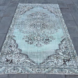 Puede incluir: Una alfombra rectangular de estilo vintage con un diseño de medallón central. La alfombra presenta un aspecto descolorido y desgastado con un fondo azul claro y patrones intrincados en tonos grises y negros. El borde de la alfombra tiene un diseño detallado y repetitivo.