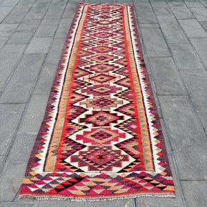 Könnte beinhalten: Ein langer, rechteckiger Läuferteppich mit geometrischem Muster. Der Teppich hat ein zentrales Design aus Rauten und Dreiecken in Rot-, Orange-, Lila- und Weißtönen. Die Ränder haben einen Zickzack-Rand. Der Teppich liegt auf einer grauen Steinoberfläche.