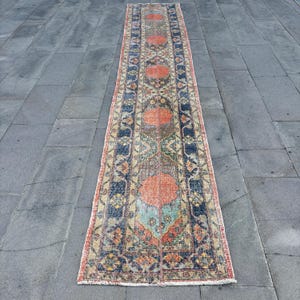 May include: A long, rectangular rug with an intricate pattern. The rug features a central design of orange medallions, surrounded by blue, green, and beige geometric shapes. The edges are bordered with a red hue, and the rug appears to be made of a woven material.