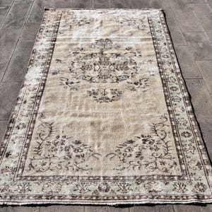 May include: A rectangular, vintage rug with a beige central field and a dark brown border. The rug features a central floral medallion and corner floral designs. The border has a repeating pattern in brown and green.