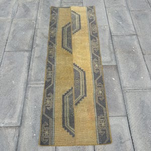 Puede incluir: Una alfombra rectangular de estilo vintage con un campo central de color amarillo mostaza y bordes geométricos y un diseño central de color gris oscuro. La alfombra probablemente está hecha de material tejido y está diseñada para un pasillo o entrada.
