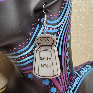 May include: White salt shaker shaped earrings with the text "Salty Bitch" on the front. The earrings are hanging from a silver hook.
