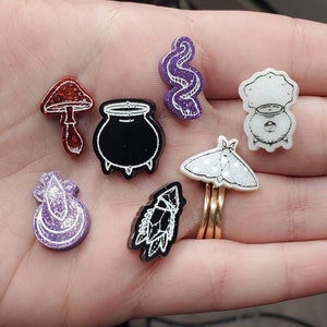 May include: A set of seven resin pins featuring various witchy and magical themes. The pins are white, black, purple, and red with white line drawings. The designs include a cauldron, a mushroom, a moth, a snake, a crystal, a crescent moon, and a potion bottle.