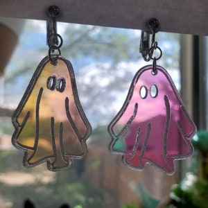May include: A pair of iridescent ghost-shaped earrings. The earrings are made of clear plastic with a holographic finish. One earring is a light pink color and the other is a light yellow color.
