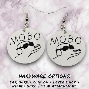 May include: Pair of white, circular earrings featuring a black and white dog wearing sunglasses, with the word "MOBO" above the dog. The earrings have silver ear wires. Text at the bottom reads "HARDWARE OPTIONS: EAR WIRE / CLIP ON / LEVER BACK / KIDNEY WIRE / STUD ATTACHMENT."