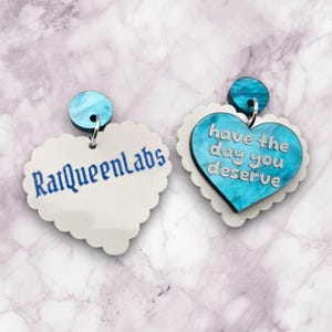 Pode incluir: Um par de brincos em forma de cora&ccedil;&atilde;o com um design de m&aacute;rmore azul e branco. Um brinco tem o texto "have the day you deserve" e o outro brinco tem o texto "RalQueenLabs".