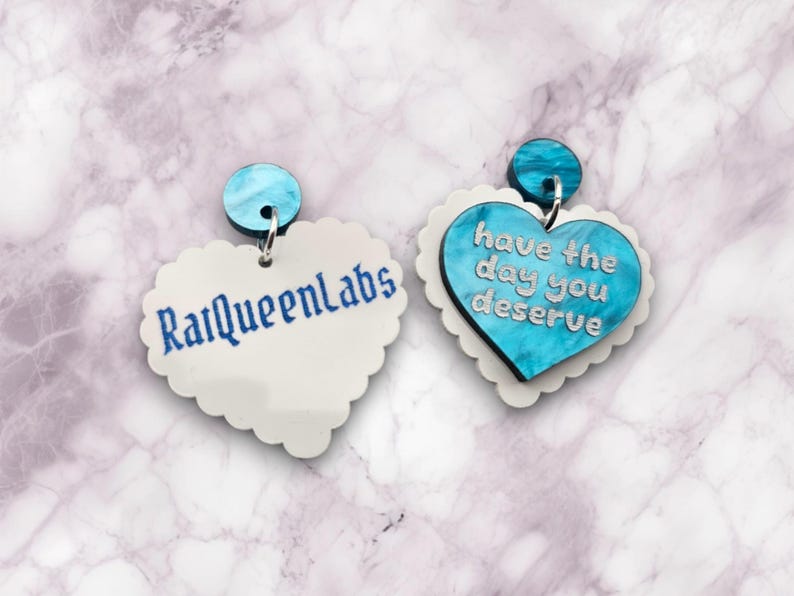 Pode incluir: Um par de brincos em forma de cora&ccedil;&atilde;o com um design de m&aacute;rmore azul e branco. Um brinco tem o texto "have the day you deserve" e o outro brinco tem o texto "RalQueenLabs".