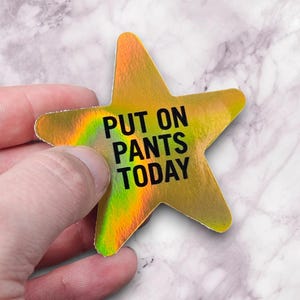 May include: A gold star-shaped sticker with the text "PUT ON PANTS TODAY" in black. The sticker has a holographic effect with rainbow colors visible. The sticker is held by a hand against a marble background.
