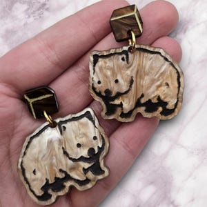 May include: A pair of wombat-shaped earrings. The earrings feature a light brown and cream marbled design with black outlines. The top of each earring is a dark brown square. The earrings are connected with a gold-colored hook.