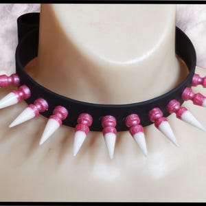 K&ouml;nnte beinhalten: Eine schwarze Choker-Halskette mit pinkfarbenen Glitzerakzenten und wei&szlig;en Spikes. Die Spikes sind in einem Kreis um den Choker angeordnet.