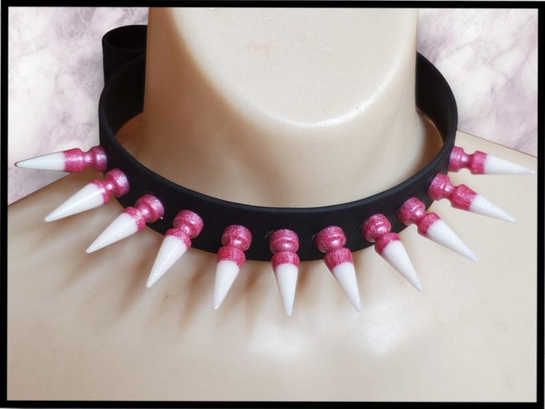 K&ouml;nnte beinhalten: Eine schwarze Choker-Halskette mit pinkfarbenen Glitzerakzenten und wei&szlig;en Spikes. Die Spikes sind in einem Kreis um den Choker angeordnet.