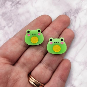 May include: Two green frog-shaped charms with pink hearts on their cheeks and a yellow circle in the center. The charms are made of resin and are about 1 inch in size.