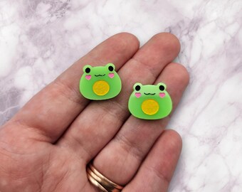 May include: Two green frog-shaped charms with pink hearts on their cheeks and a yellow circle in the center. The charms are made of resin and are about 1 inch in size.