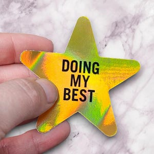 May include: A holographic star-shaped sticker with the text "DOING MY BEST" in black. The sticker has a yellow and green iridescent finish. The sticker is held in a hand.