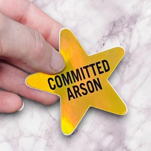 May include: A holographic star-shaped sticker with the words "COMMITTED ARSON" in black text. The sticker is held by a hand against a marble background. The sticker has a yellow and orange iridescent finish.