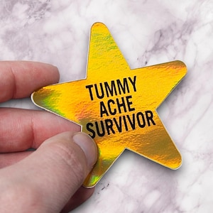 May include: A gold star-shaped sticker with the text "TUMMY ACHE SURVIVOR" in black. The sticker has a holographic finish, creating a rainbow effect. The sticker is held by a hand against a marble background.