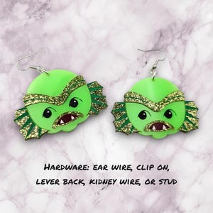 May include: Green and gold creature from the black lagoon earrings. The earrings are made of acrylic and have a glitter finish. The earrings are available in a variety of hardware options, including ear wire, clip on, lever back, kidney wire, or stud.
