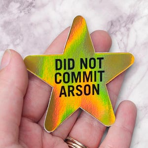 Puede incluir: Una pegatina holográfica en forma de estrella con el texto "DID NOT COMMIT ARSON" en negro. La pegatina tiene un esquema de color amarillo, verde y naranja. La pegatina está sujeta en una mano.