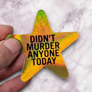 May include: A star-shaped sticker with a holographic finish in shades of gold, green, and orange. The sticker has the text "DIDN'T MURDER ANYONE TODAY" in bold black letters. The sticker is held by a hand.