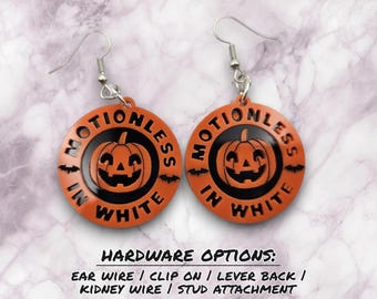 May include: A pair of round orange and black earrings featuring a central jack-o'-lantern graphic, small bat silhouettes, and the text MOTIONLESS IN WHITE. The earrings include silver-toned ear wires for attachment to the earlobes.