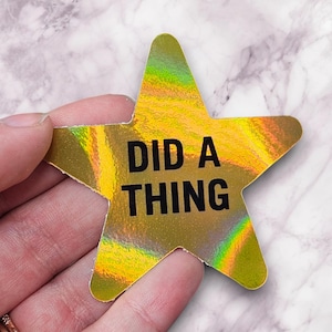 May include: A holographic gold star-shaped sticker with the text "DID A THING" in black. The sticker has a rainbow sheen and is held by a hand.