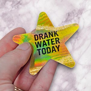 May include: A star-shaped sticker with a holographic, yellow, and green surface. The sticker has the words "DRANK WATER TODAY" in black block letters. The sticker is held in a person's hand.