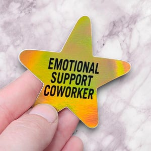 May include: A star-shaped sticker with the text "EMOTIONAL SUPPORT COWORKER" in black. The sticker has a gradient of yellow, orange, and green colors. The sticker is held by a hand against a marble background.