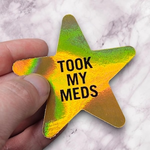 May include: A holographic star-shaped sticker with the text "TOOK MY MEDS" in black. The sticker has a gradient of colors, including gold, green, and orange. The sticker is held by a hand.