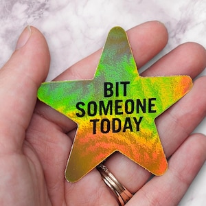 May include: A holographic star-shaped sticker with the text "BIT SOMEONE TODAY" in black. The sticker has a gradient of green, yellow, and orange colors. The sticker is held in a hand.