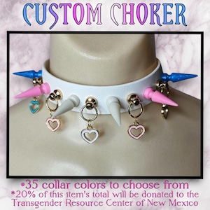 May include: White leather choker with silver spikes and pink and blue charms. The text "CUSTOM CHOKER" is at the top of the image. The text "•35 collar colours to choose from *20% of this item's total will be donated to the Transgender Resource Center of New Mexico" is at the bottom of the image.