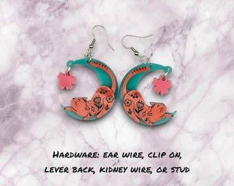 May include: A pair of earrings featuring a crescent moon design with a sleeping cat and a pink flower charm. The earrings are available in a variety of hardware options, including ear wire, clip on, lever back, kidney wire, or stud.