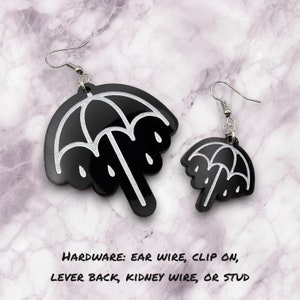 May include: Black umbrella-shaped earrings with white outlines and dangling raindrops. The earrings are in two sizes and have silver ear wires. The text at the bottom says "HARDWARE: EAR WIRE, CLIP ON, LEVER BACK, KIDNEY WIRE, OR STUD."