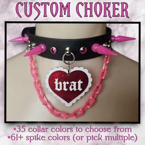 May include: A black choker with pink spikes and a pink chain. The chain has a heart-shaped charm with the word "brat" written on it.