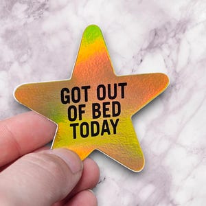 May include: A star-shaped sticker with a gradient of yellow, orange, and green. The sticker has the text "GOT OUT OF BED TODAY" in black. The sticker is held by a hand against a marble background.