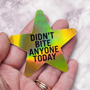 May include: A holographic star-shaped sticker with the text "DIDN'T BITE ANYONE TODAY" in black. The sticker has a gradient of green, yellow, and gold colors. The sticker is held in a person's hand.