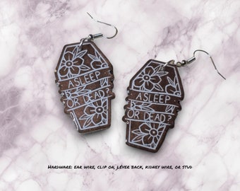 May include: A pair of coffin-shaped earrings with the text "ASLEEP OR DEAD?" in white lettering on a brown background. The earrings are decorated with white floral designs.