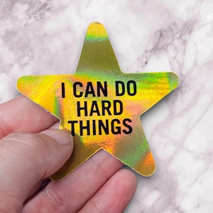 May include: A holographic star-shaped sticker with the text "I CAN DO HARD THINGS" in black. The sticker has a shiny, iridescent finish with yellow, green, and orange hues. The sticker is held by a hand.