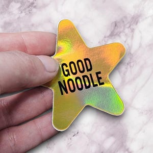 May include: A holographic star-shaped sticker with the words "GOOD NOODLE" in black. The sticker has a shiny, iridescent finish with yellow, green, and orange hues. The sticker is held against a marble background.