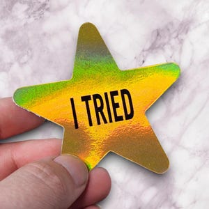 May include: A holographic star-shaped sticker with the words "I TRIED" in black. The sticker has a gold and green iridescent finish. The sticker is held by a hand against a marble background.