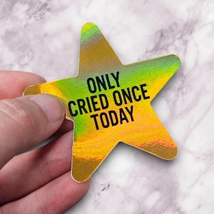 May include: A holographic star-shaped sticker with the text "ONLY CRIED ONCE TODAY" in black. The sticker has a gradient of yellow, green, and gold colors. The sticker is held by a hand against a marble background.