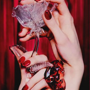 May include: A clear glass rose sculpture is held in front of a red curtain. The sculpture is held by a person with red nail polish and a large red gemstone ring. The rose has detailed petals and a long stem.