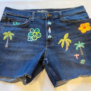 May include: Blue denim shorts with a raw hem, featuring hand-painted tropical designs. The shorts are adorned with colorful palm trees, flowers, and other tropical motifs. The brand name "Goodfellow" is visible on the waistband.