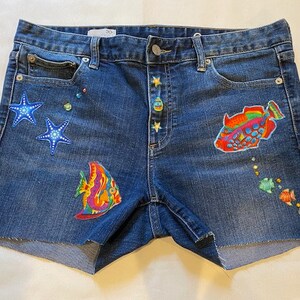May include: Blue denim shorts with embroidered sea life designs, including starfish, fish, and a pufferfish. The shorts have a button fly and five pockets.
