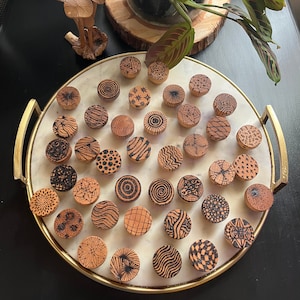 Puede incluir: Una bandeja de 25 pomos de madera con varios patrones en blanco y negro. Los pomos están dispuestos en un patrón circular en una bandeja de mármol blanco con un borde dorado.