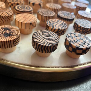 K&ouml;nnte beinhalten: Eine Sammlung von Holzkn&ouml;pfen mit verschiedenen geometrischen Mustern in Schwarz und Wei&szlig;. Die Kn&ouml;pfe sind in einem Kreis auf einer wei&szlig;en Marmorfl&auml;che mit einem goldenen Rand angeordnet.